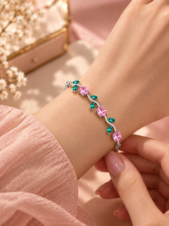 A close-up of a delicate silver bracelet with pink and green gemstones, worn on a woman's wrist with a soft pink sleeve visible.