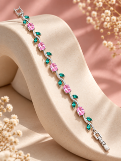 A silver-toned bracelet with alternating pink and green teardrop gemstones, displayed on a beige sculptural stand against a soft pink background with dried floral accents.