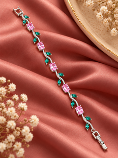 A silver-toned bracelet with alternating pink and green gemstones, styled on draped pink satin fabric with dried white flowers and a gold-rimmed plate in the background.