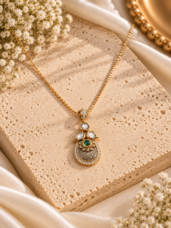 A gold pendant necklace with a detailed traditional Indian design, featuring a green stone and white stones, displayed on a textured beige stone block with cream satin fabric and dried flowers.