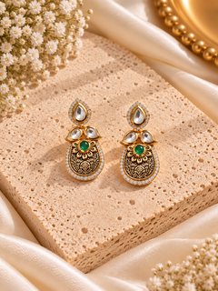 A pair of ornate gold-toned traditional Indian jhumka earrings with green stones and clear stones, displayed on a textured beige stone block with cream satin fabric and dried flowers.