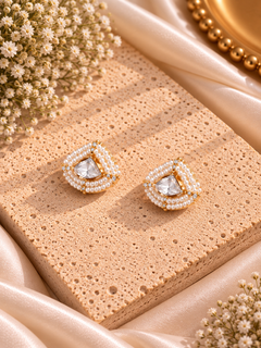 A pair of gold and pearl stud earrings with clear heart-shaped crystals, displayed on a textured beige stone slab with draped satin fabric and baby's breath flowers.
