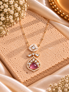 A gold-tone pendant necklace with a large pink teardrop-shaped stone and clear rhinestone accents, displayed on a textured beige stone slab with satin fabric and dried flowers.