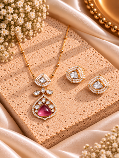 Gold-toned jewelry set with a pendant necklace and matching earrings, featuring clear stones and a prominent pink gemstone, styled on a textured beige stone slab with satin fabric and dried flowers. demi fine jewellery • demi fine jewellery india • polki jewellery • polki jewellery set • jewellery set • ethnic jewellery • ethnic jewellery set • festive jewellery • festive jewellery set • occasion jewellery • wedding jewellery • traditional jewellery • jewellery for saree • simple jewellery for saree • moder