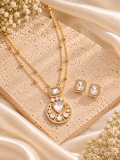 Gold-toned jewelry set with a layered necklace and matching rectangular stud earrings, styled on a beige stone slab with cream satin fabric and small white flowers.
