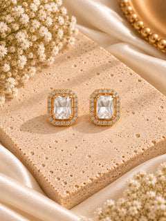 A pair of gold-toned rectangular stud earrings with clear cushion-cut center stones and small clear stones around the halo, displayed on a beige textured stone slab with cream satin fabric and dried flowers.