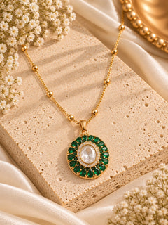 A gold pendant necklace with a circular green gemstone halo and a central clear oval stone, displayed on a textured beige stone slab with cream satin fabric and small white flowers.