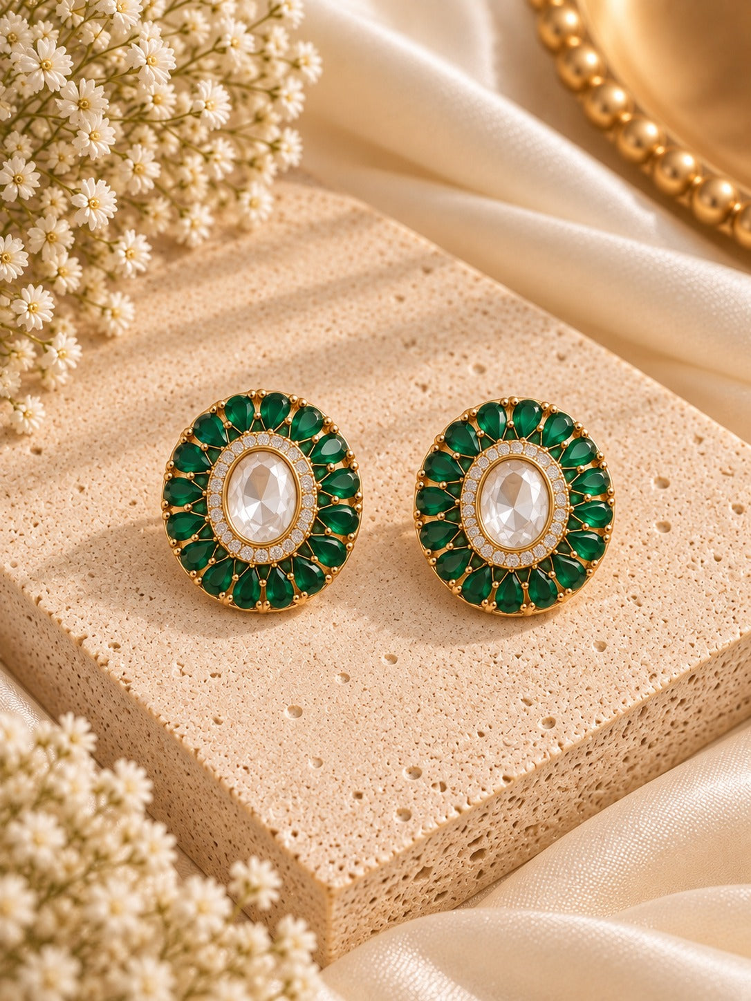A pair of oval gold-tone earrings with a central white marquise-cut stone surrounded by a halo of green stones and small clear accents, displayed on a beige stone slab with cream fabric and dried flowers.