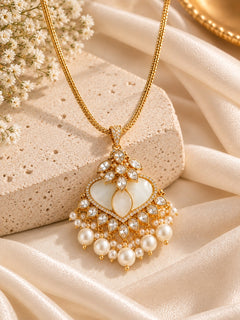 An ornate gold pendant necklace with a heart-shaped white inlay, surrounded by small pearls and clear rhinestones, displayed on draped cream satin fabric with a stone prop.