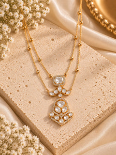 A gold-tone double-strand necklace with a detailed pendant set with clear stones, displayed on a textured beige stone slab with satin fabric and dried flowers.