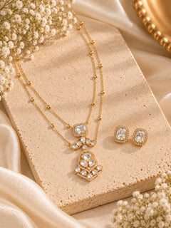 Gold-tone jewelry set featuring a double-strand necklace with a floral pendant and matching stud earrings, styled on a beige stone slab with cream satin fabric and small white flowers.