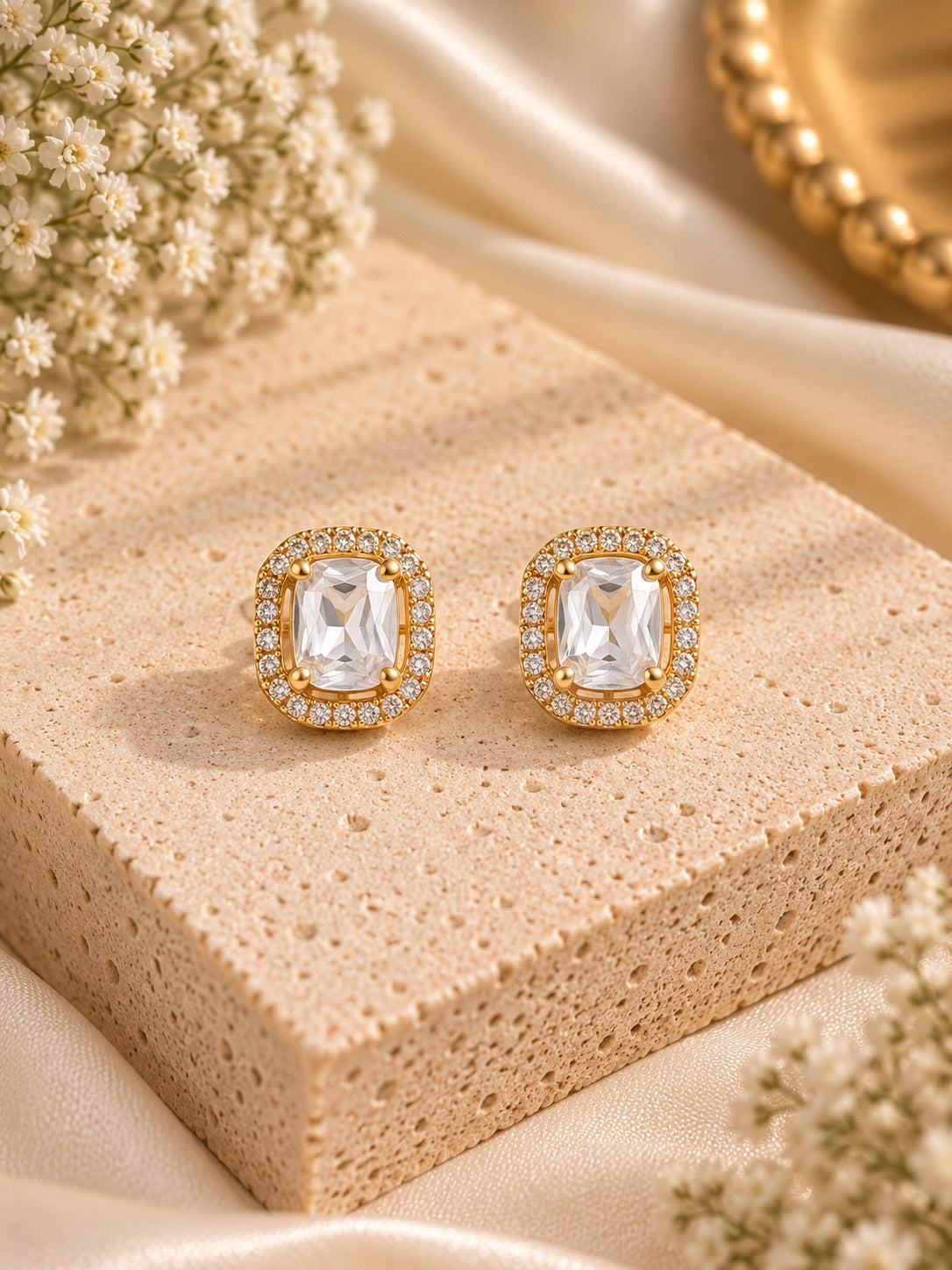 A pair of gold-toned rectangular stud earrings with clear cushion-cut center stones and a halo of small clear stones, displayed on a beige textured block with cream flowers and satin fabric.