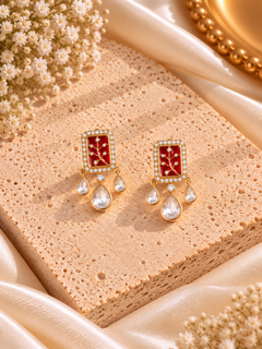A pair of gold-toned statement earrings with rectangular red enamel panels featuring a gold floral motif, accented with small clear stones and dangling teardrop-shaped clear stones, displayed on a textured beige stone block with satin fabric and dried flowers.