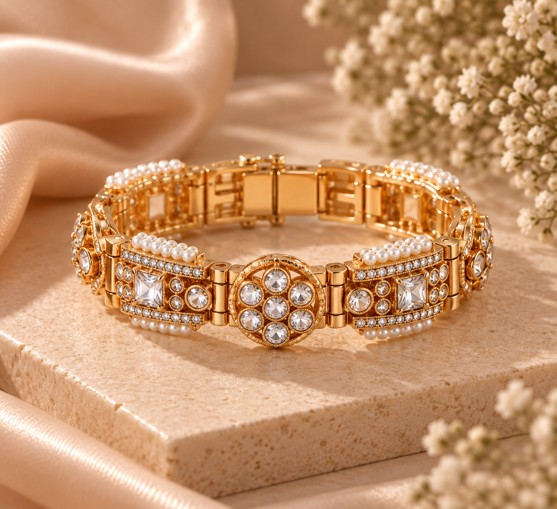 A gold-toned bracelet with multiple rows of clear rhinestones and faux pearls, displayed on a beige stone slab with soft satin fabric and dried flowers in the background.