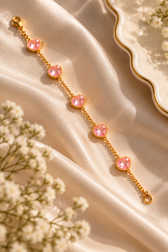 A gold chain bracelet with five pink heart-shaped crystals, styled on draped cream satin fabric with white baby's breath flowers.