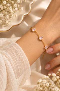 A delicate gold chain bracelet with three pink heart-shaped crystals, shown worn on a woman's wrist. The background features soft white fabric, white baby's breath flowers, and a gold-rimmed dish.