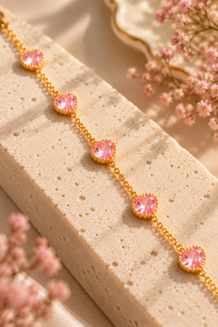 A gold chain bracelet with five pink heart-shaped gemstones, styled on a textured beige stone block with soft pink flowers in the background.