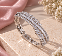 A silver-toned hinged bangle bracelet with three parallel rows of baguette-cut clear stones and leaf-shaped clear stones on the outer band, displayed on a beige stone slab with pink satin and dried flowers in the background.