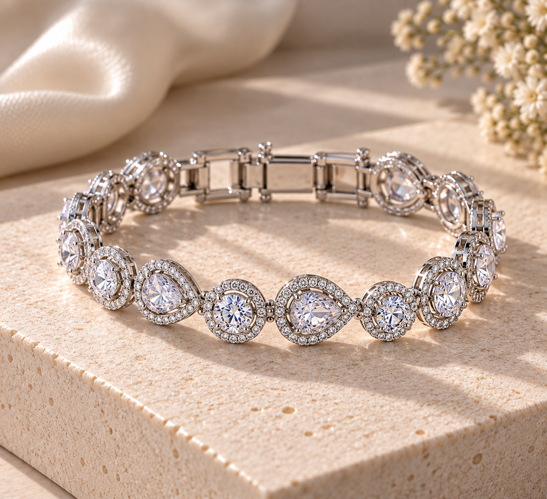 A silver-toned tennis bracelet with round and marquise-cut clear stones in halo settings, displayed on a textured beige stone surface with soft cream fabric and dried flowers in the background.