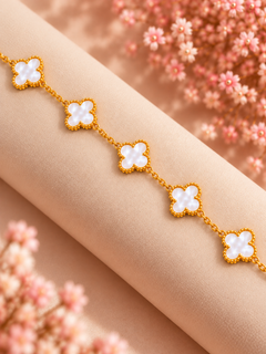 A gold chain bracelet with five white clover-shaped links, displayed on a beige fabric roll with soft pink floral decorations in the background.