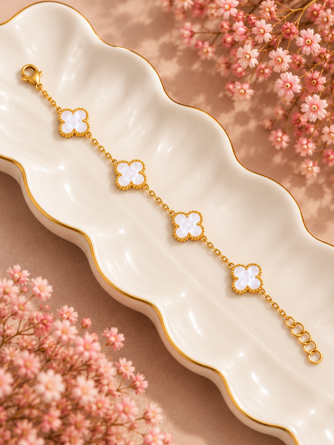 A gold chain bracelet with four white flower charms, displayed on a white scalloped dish with gold trim, surrounded by pink flowers.