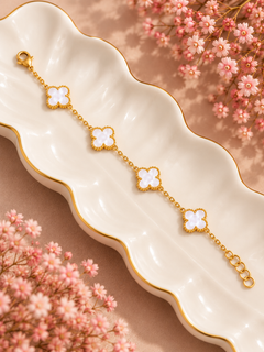 A gold chain bracelet with four white flower charms, displayed on a white scalloped dish with gold trim, surrounded by pink flowers.