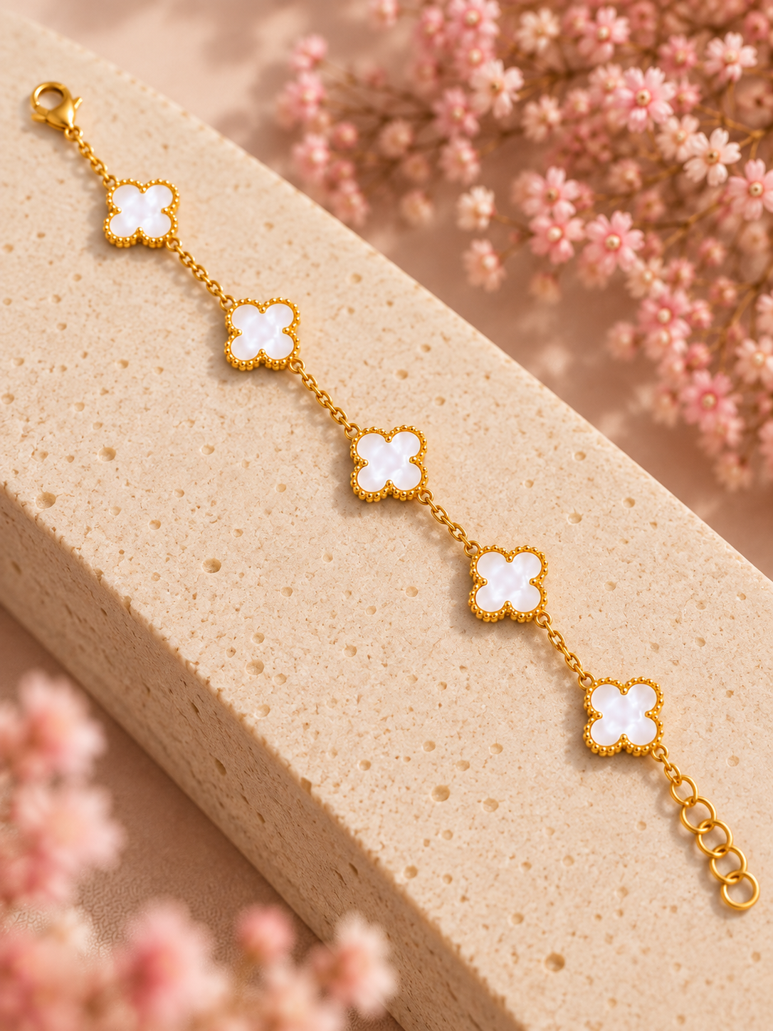 A gold chain bracelet with five white clover-shaped charms, displayed diagonally on a textured beige stone slab with soft pink flowers in the background.