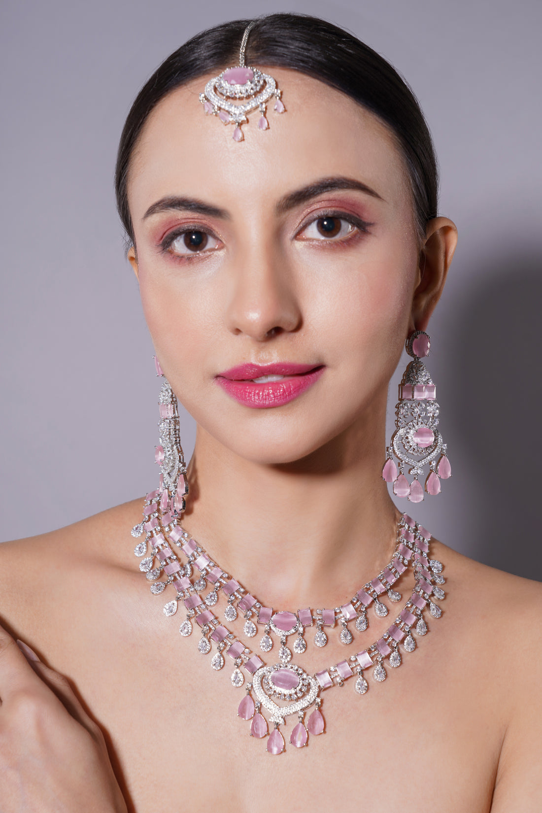 Woman wearing a pink and silver jewelry set including necklace, earrings, and headpiece against a gray background