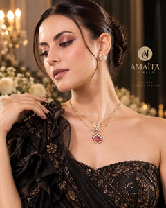 A woman wearing a gold-tone double-layered necklace with a pinkish-red teardrop-shaped pendant and matching stud earrings, styled with a black embellished dress in an elegant indoor setting. The brand logo 'AMAITA JEWELLERY' is visible on the right.