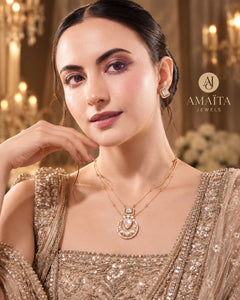 A woman wearing a gold-tone necklace and matching stud earring with a pearl-like center, styled with a sequined beige gown in an elegant indoor setting. The brand name 'AMAITA JEWELS' is visible in the upper right.