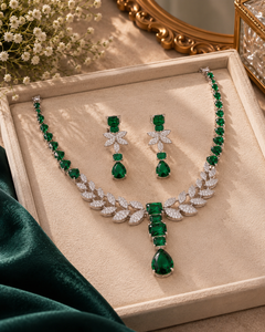 A green gemstone jewelry set displayed on a beige velvet jewelry box, featuring a necklace and matching earrings with emerald-cut and round green stones set in silver-toned metal, accented with clear crystals. The set is styled with a dark green velvet cloth and baby's breath flowers.
