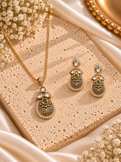 Gold-toned traditional jewelry set featuring a necklace and matching earrings with ornate filigree detailing, green stone accents, and clear stones, arranged on a textured beige stone slab with cream satin fabric and dried flowers.