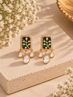 A pair of gold-plated drop earrings with rectangular green enamel panels featuring a tree motif set with small clear stones, and teardrop-shaped clear crystal drops below, displayed on a beige stone block with baby's breath flowers.
