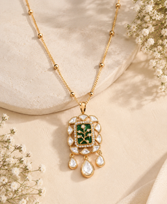 Gold pendant necklace with a green enamel floral center and clear teardrop stones, styled on a beige fabric surface with small white flowers.