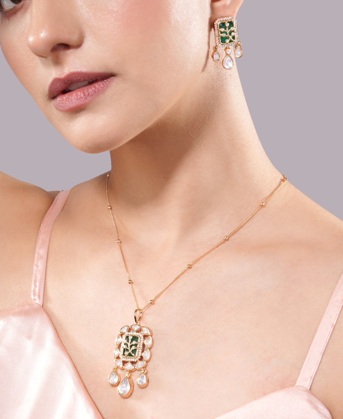 Close-up of a woman wearing a gold necklace and earrings with green gemstones on a gray background. jewellery set, pendant with chain, pendant set, pendants