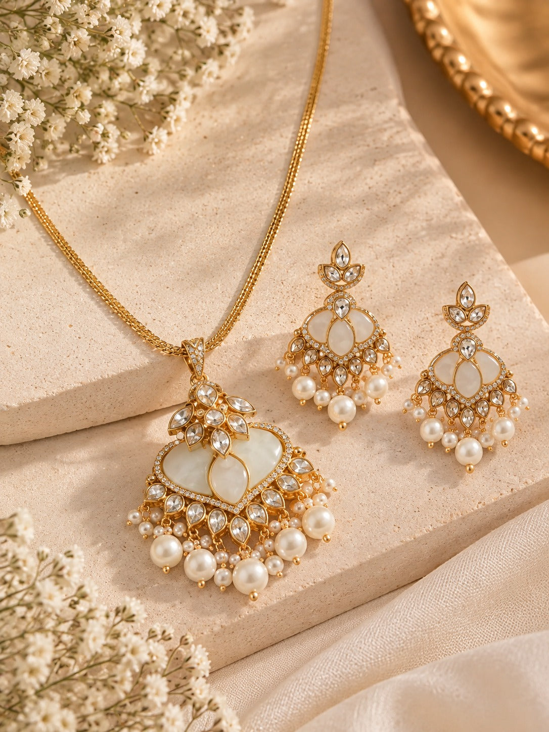 Gold-toned traditional jewelry set featuring a pendant necklace and matching earrings with white pearl-like beads, clear rhinestone embellishments, and a soft beige textured background with floral props.