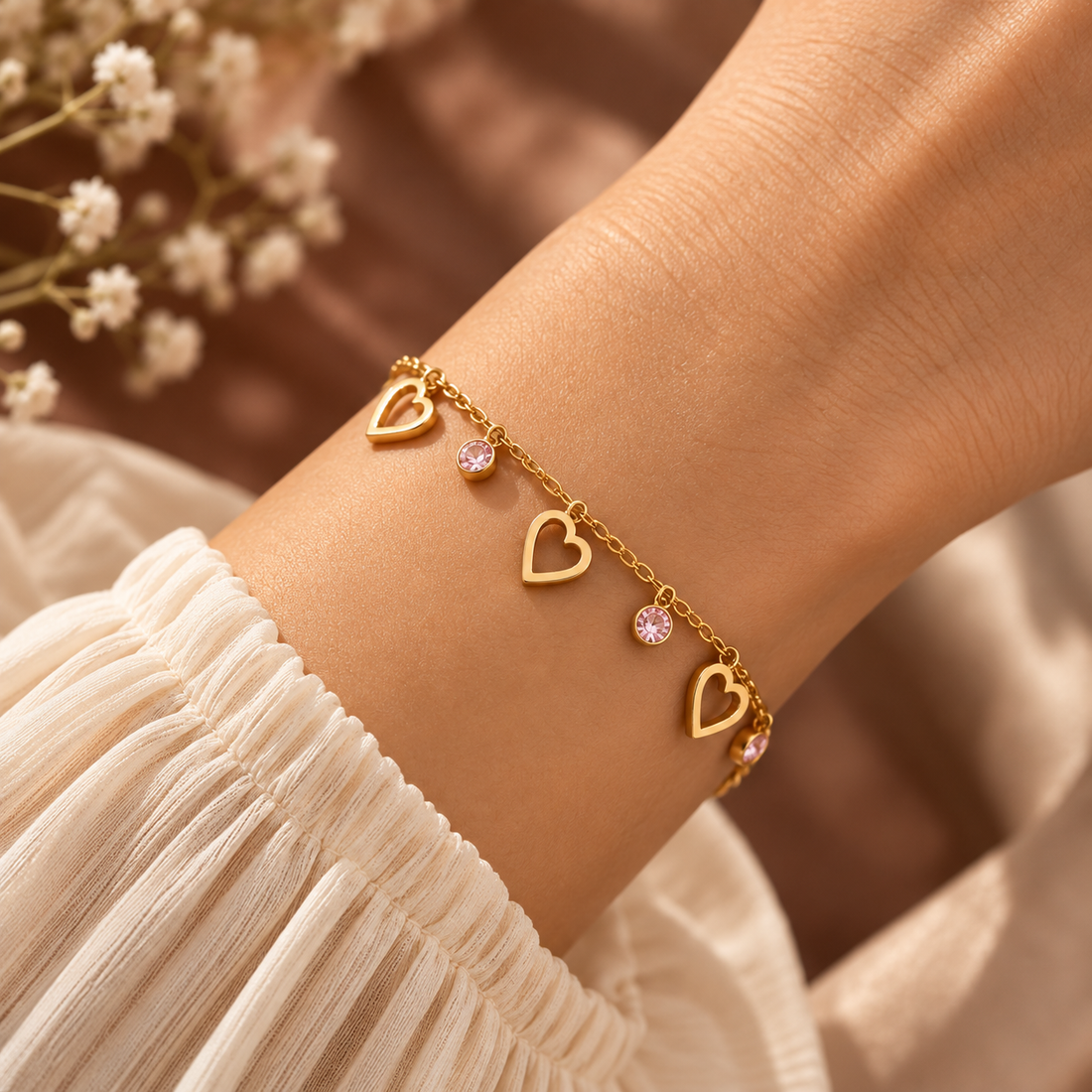 A delicate gold chain bracelet with three open heart charms and two small pink crystal accents, shown worn on a person's wrist. The person is wearing a white pleated blouse, with soft, blurred baby's breath flowers in the background.