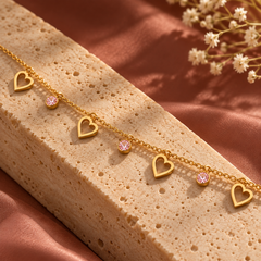 A gold chain bracelet with five dangling heart charms and two small round pink gemstones, displayed on a textured beige stone slab over a warm pink satin fabric background.