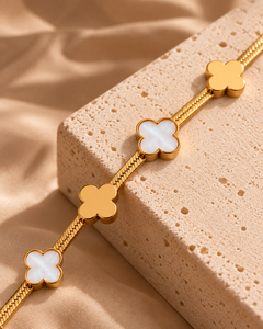 Gold-tone chain bracelet with four white clover-shaped charms, displayed diagonally on a textured beige stone block against a draped beige satin fabric background.