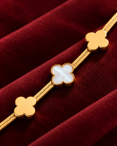 A gold-toned bracelet with three clover-shaped charms, featuring a central white mother-of-pearl clover and two gold clover charms on either end, displayed on a deep red velvet surface.