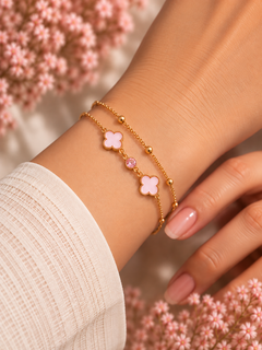 A close-up of a person's wrist wearing two delicate gold bracelets, each featuring a light pink four-leaf clover charm and a small pink gemstone, styled with soft pink flowers in the background.
