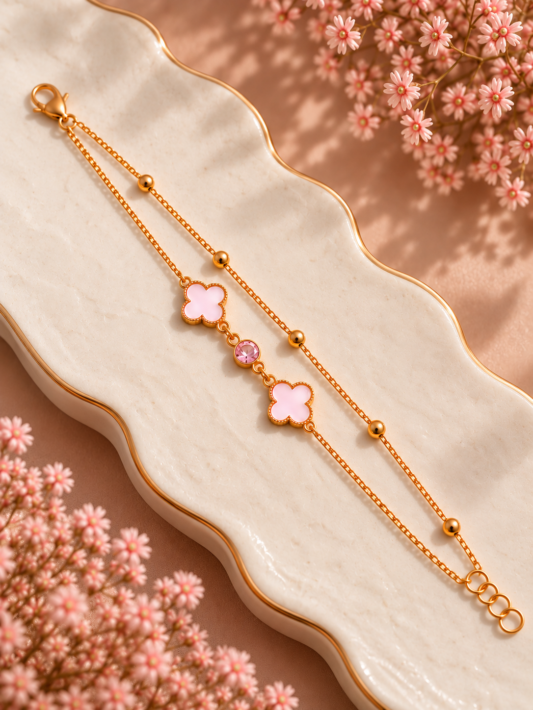 A delicate gold chain bracelet with two pink enamel four-leaf clover charms and a small pink crystal accent, displayed on a wavy cream ceramic dish with pink dried flowers.