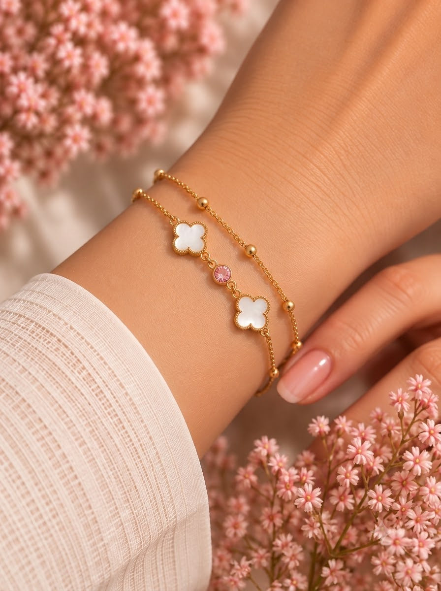 A close-up of a person's wrist wearing two delicate gold bracelets, each featuring a white four-leaf clover charm and a small pink gemstone, styled with soft pink flowers in the background.