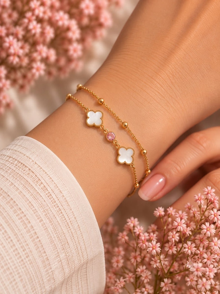 A close-up of a person's wrist wearing two delicate gold bracelets, each featuring a white four-leaf clover charm and a small pink gemstone, styled with soft pink flowers in the background.