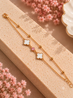 A delicate gold chain bracelet with two white clover charms and a small pink gemstone, displayed on a textured beige block with pink flowers in the background.
