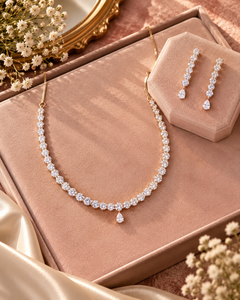 A gold-tone necklace and matching drop earrings set with clear stones, displayed on blush pink jewelry boxes with decorative dried flowers and satin fabric around them.
