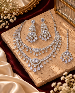 A sparkling silver-toned jewelry set arranged on a beige stone slab, featuring a layered necklace, matching earrings, and a maang tikka. The jewelry is decorated with clear teardrop and round stones and is styled on a deep red velvet fabric with cream satin and dried flowers.
