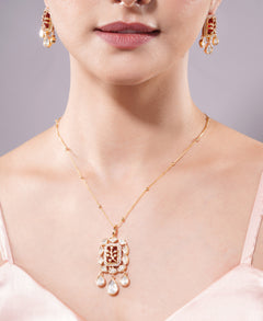 Close-up of a person wearing a gold necklace with a pendant and matching earrings on a neutral background.