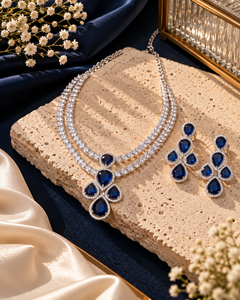 A silver-tone jewelry set with a double-strand necklace and matching earrings, featuring deep blue teardrop-shaped stones and clear crystal-like accents, styled on a beige stone slab with navy fabric, cream satin, and small white flowers.