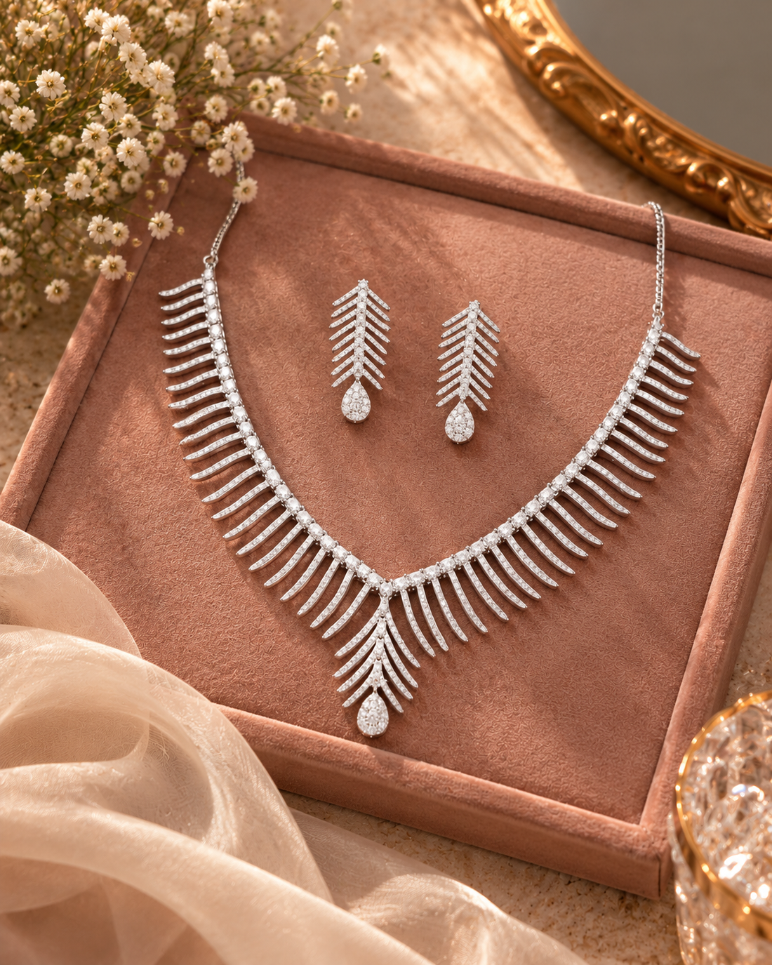A silver-toned feather-style necklace and matching drop earrings displayed on a blush pink jewelry box, with dried flowers and soft fabric props around it.